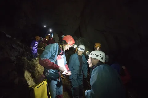 Un groupe de personnes équipées de casques et de lampes frontales participe à une exploration souterraine. À l’intérieur d’une grotte, un animateur montre un document tandis que les participant·es l’écoutent.
