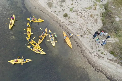 Vue aérienne de plusieurs kayaks jaunes sur une zone peu profonde, près d’une berge sableuse. Des participant·es embarquent ou débarquent tandis qu’un groupe observe depuis le rivage.