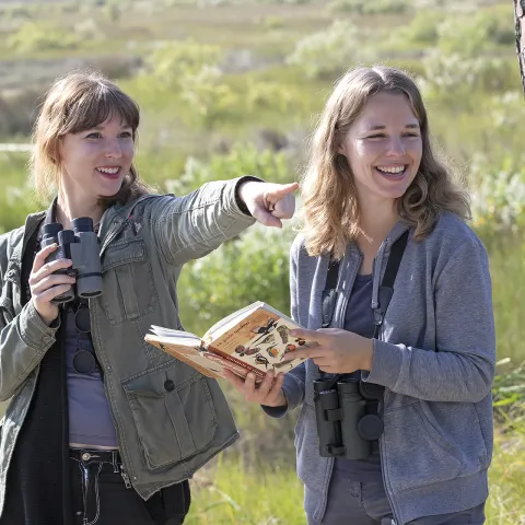 Deux jeunes femmes, équipées de jumelles, observent la nature dans un paysage de marais. L’une pointe au loin en souriant tandis que l’autre tient un guide illustré d’oiseaux