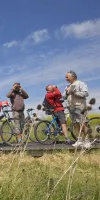 Un groupe d’adultes s’est arrêté lors d’une balade à vélo sur un chemin en pleine nature. Alignés avec leurs vélos, ils observent le paysage à l’aide de jumelles et d’appareils photo, sous un ciel bleu.